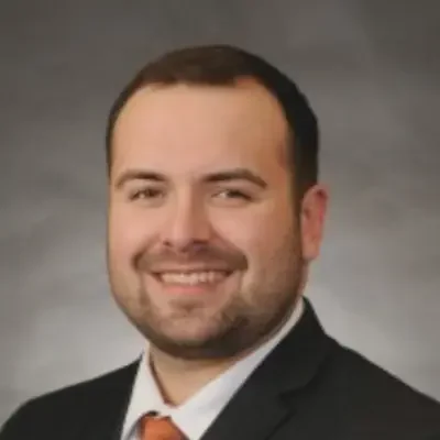 Professional headshot of Doctor Andrew smiling and wearing a dark suit, white shirt, and orange tie
